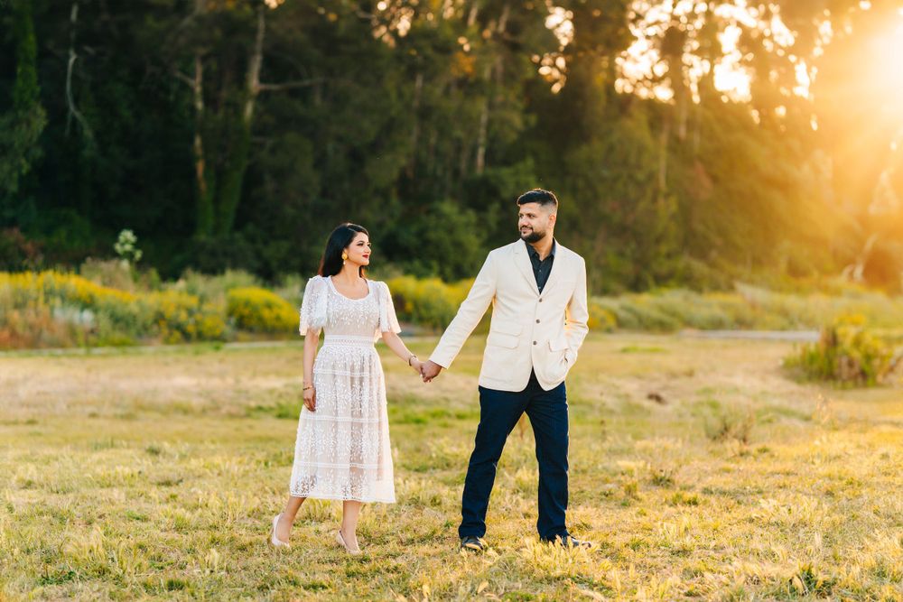 A couple in coordinated white outfits holds hands in a sunlit meadow during golden hour.