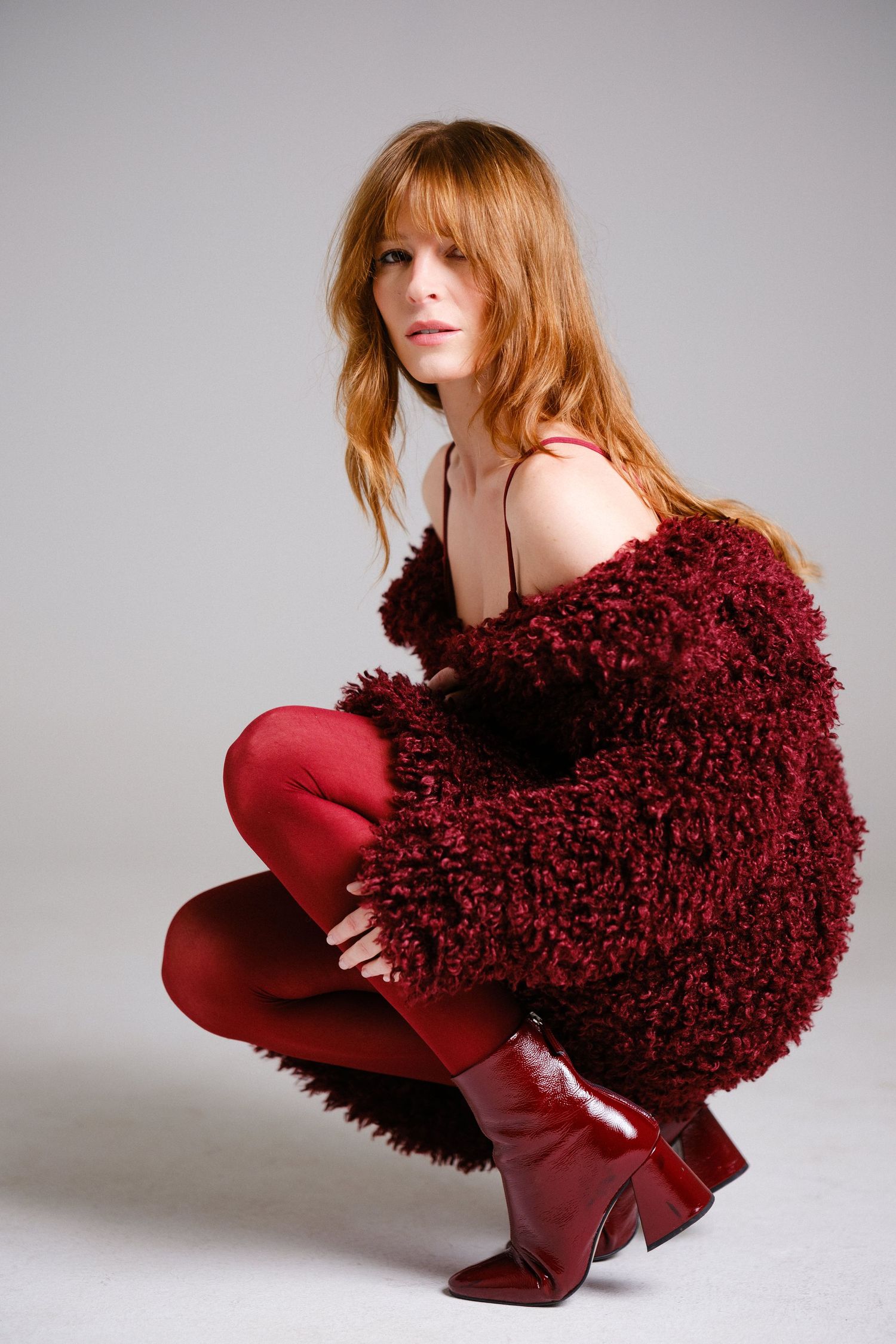 Fashion model in red tights poses with burgundy textured pillow against grey background.