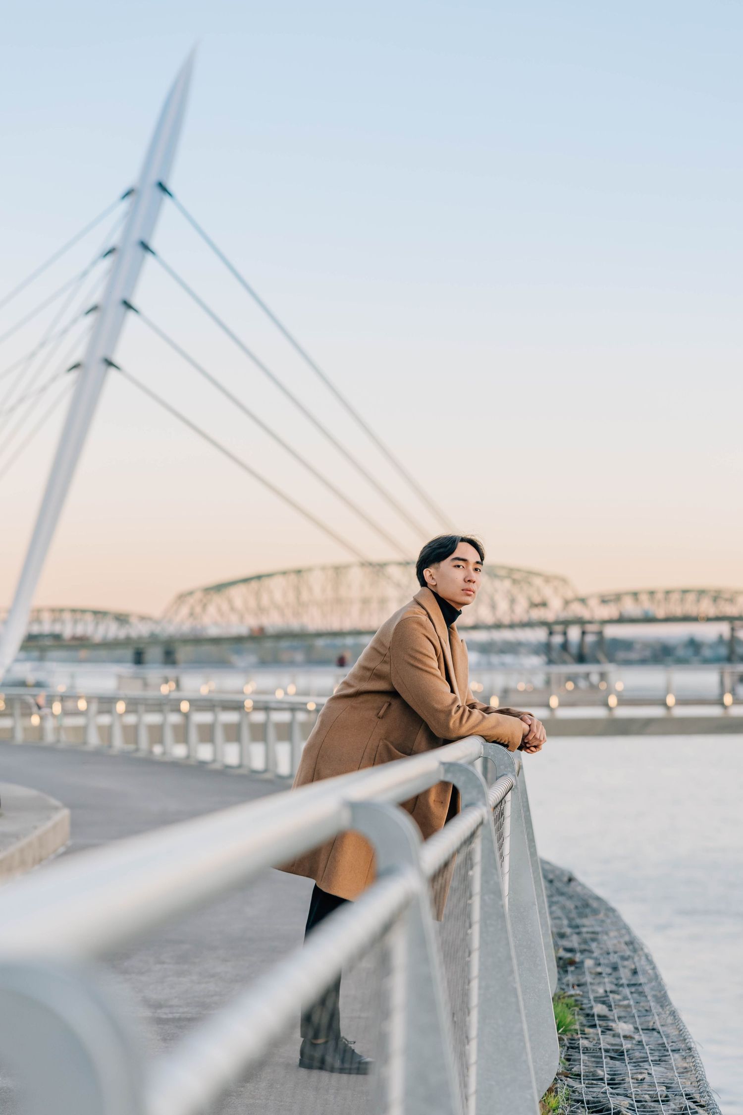 Young Asian man in camel coat leaning on bridge railing at sunset with suspension bridge in background.
