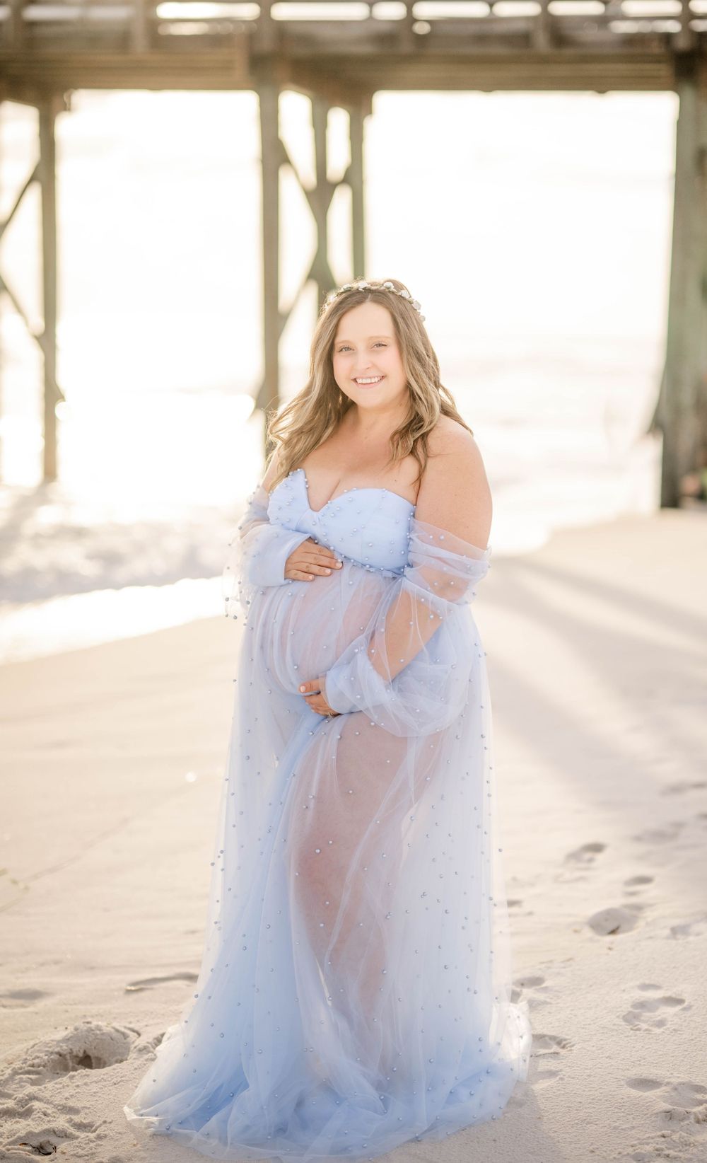Maternity beach photo featuring woman in light blue tulle gown posing by wooden pier.