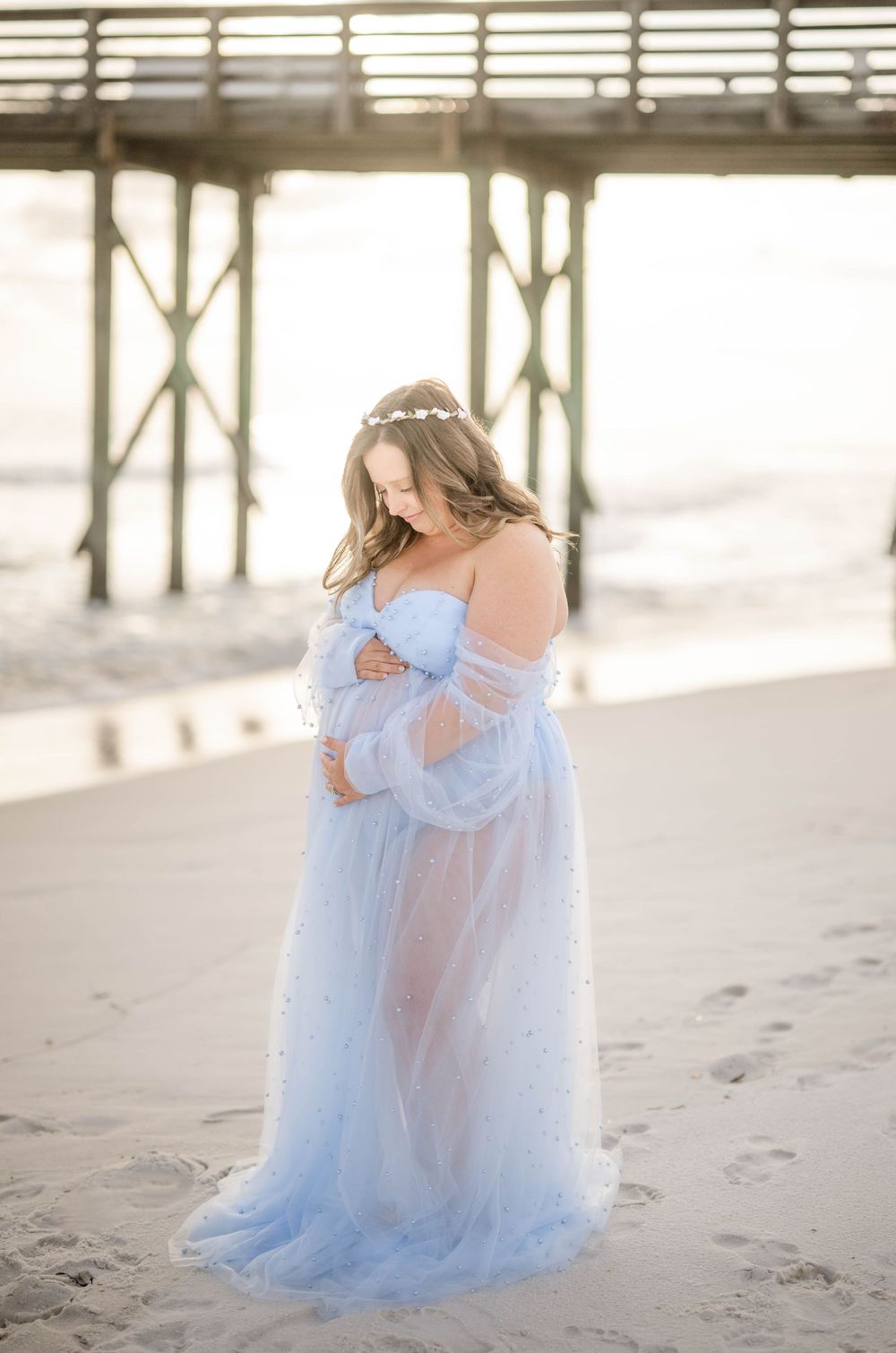 Expectant mother in flowing blue gown poses on sandy beach with wooden pier backdrop.