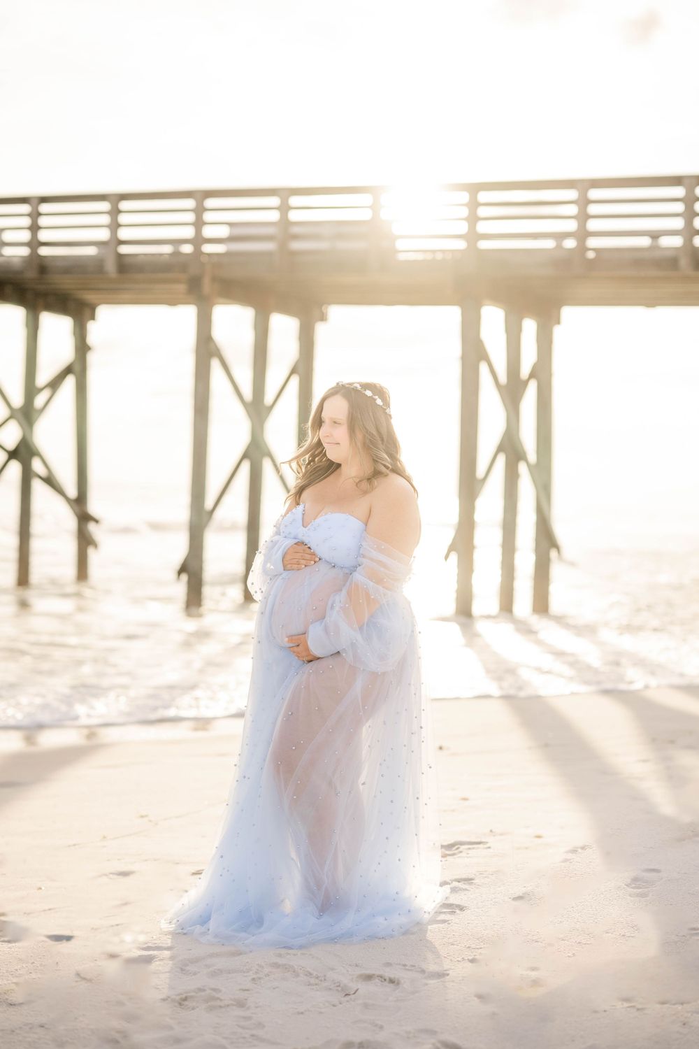 Sunlit maternity portrait with woman in sheer blue dress standing by beach pier.