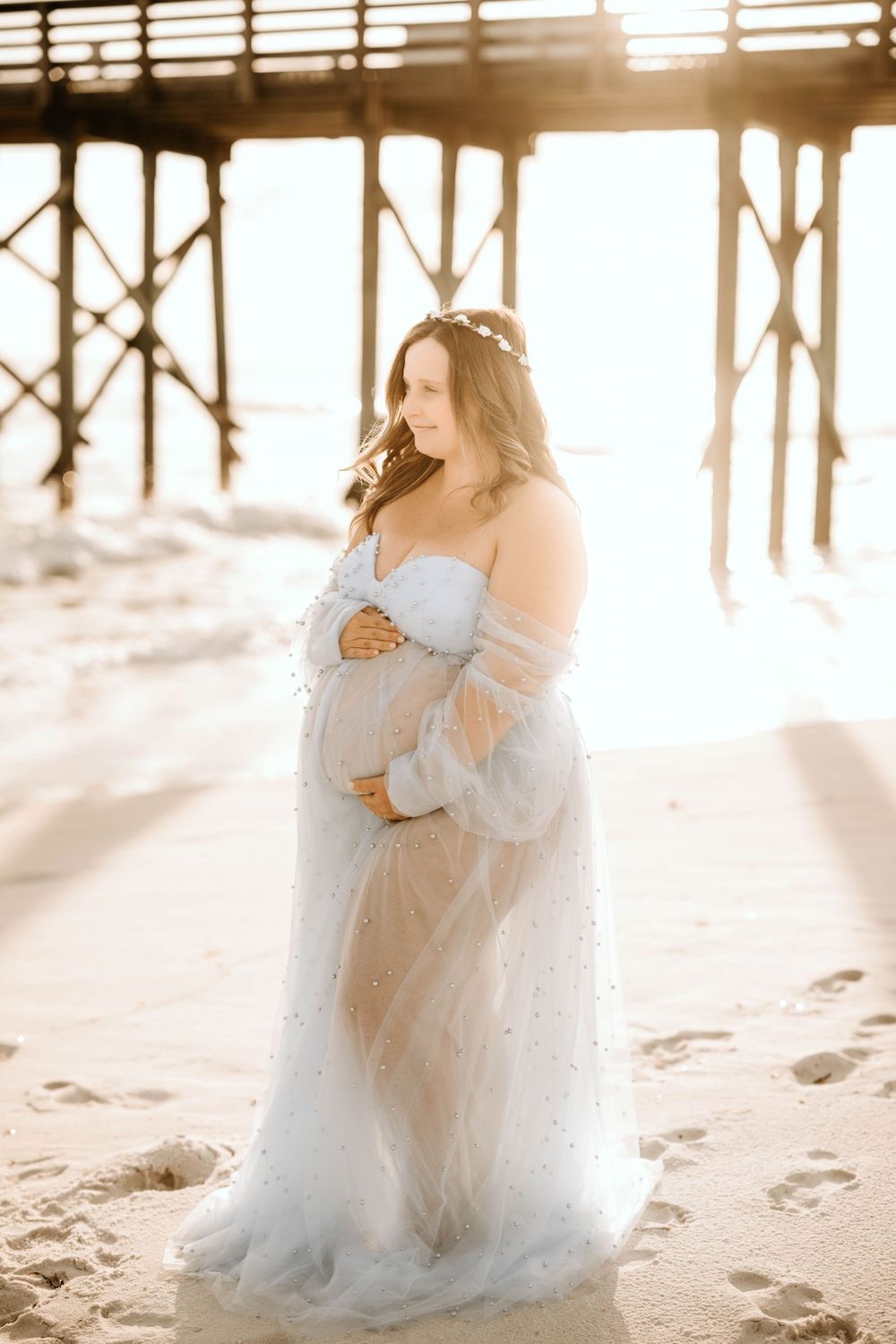 Dreamy beach maternity photo with backlit pier and flowing blue gown.