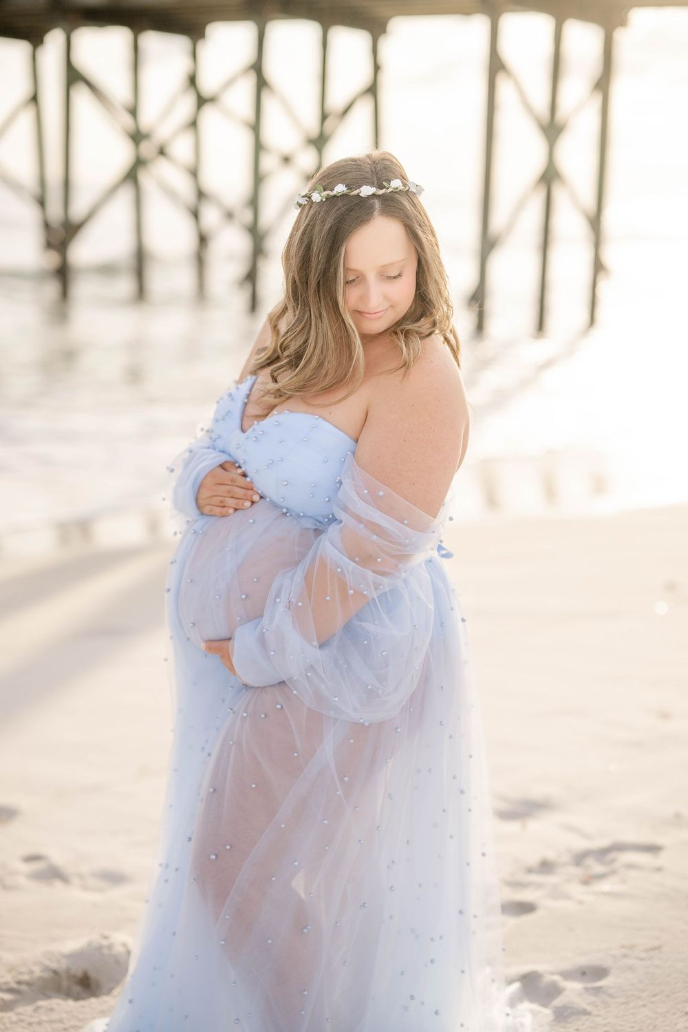 Elegant maternity portrait on beach with woman in powder blue dress and flower crown.