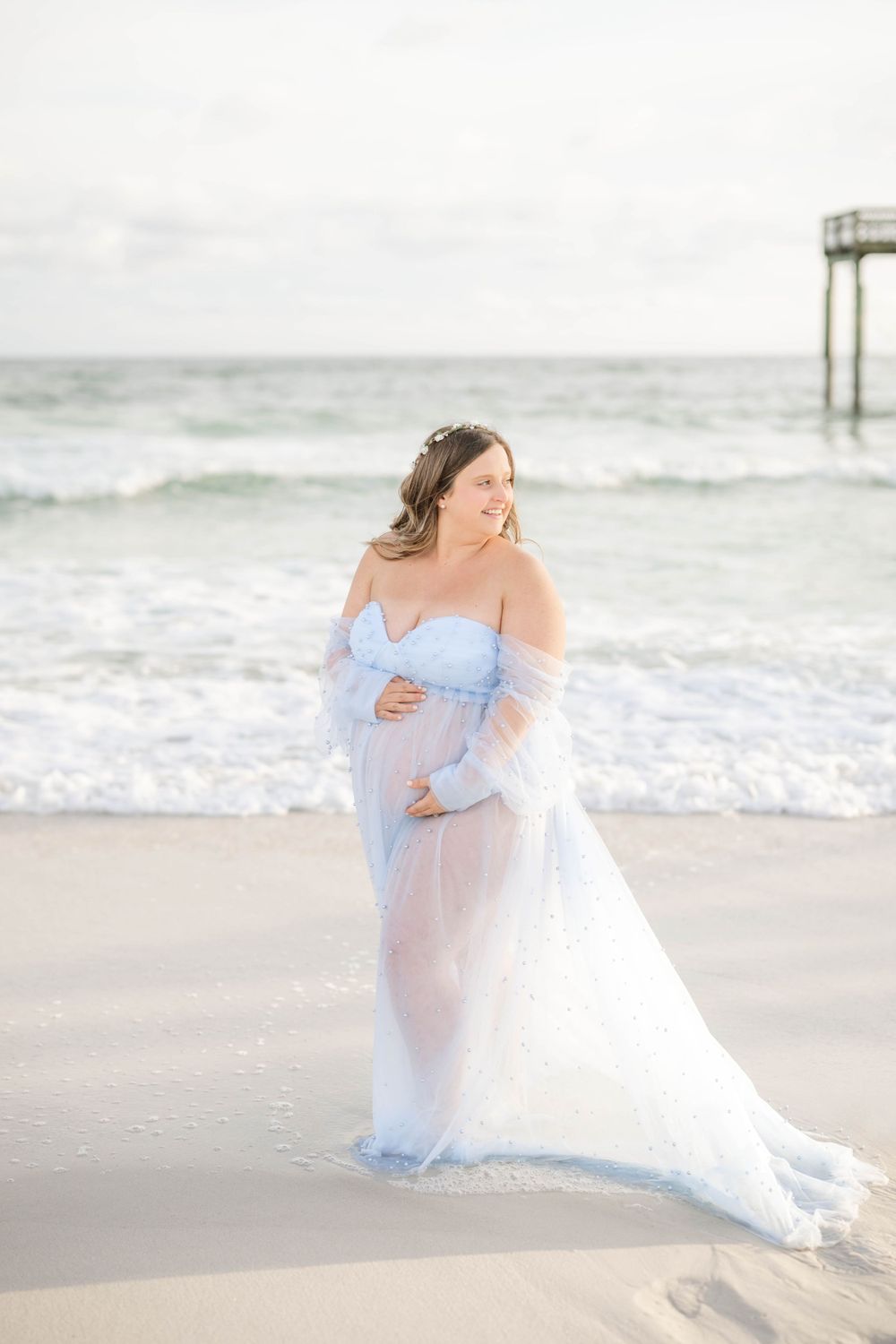 Ethereal maternity photo of an expectant mother in a flowing light blue gown standing on a beach with a pier in the distance.