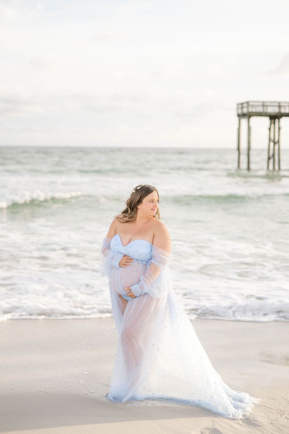Dreamy beach maternity portrait featuring a flowing blue and white dress against ocean waves and wooden pier.