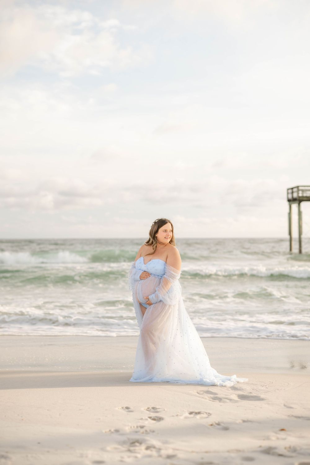 Expectant mother in an airy blue gown poses on sandy beach with footprints and pier in background.