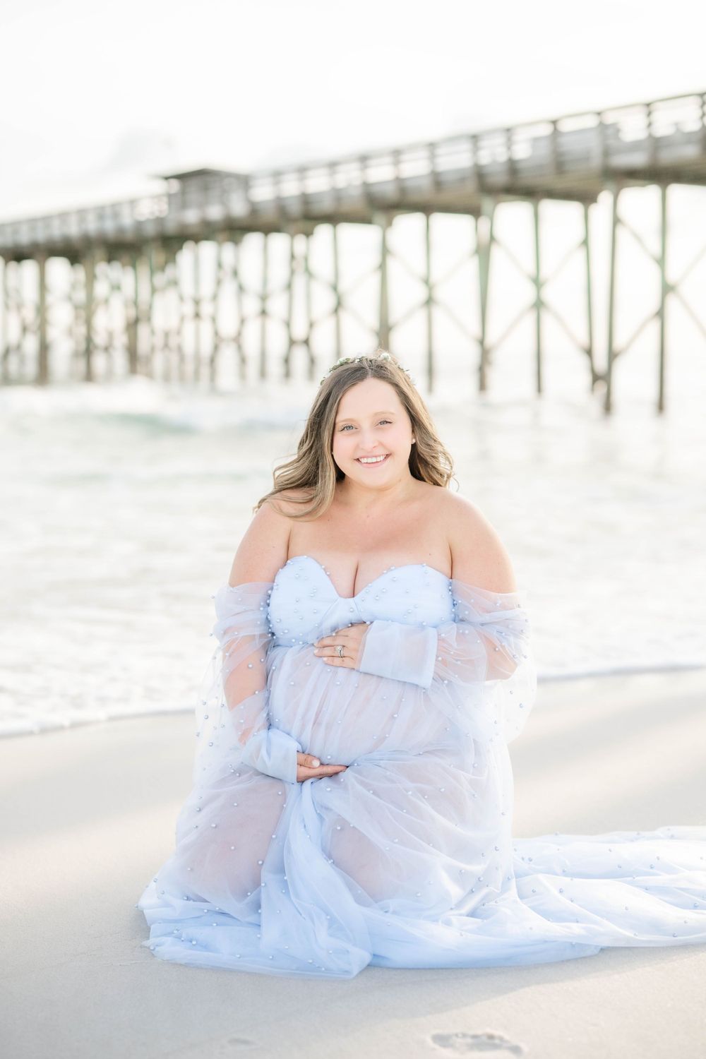 Beachside maternity photo with wooden pier backdrop and flowing off-shoulder blue gown.