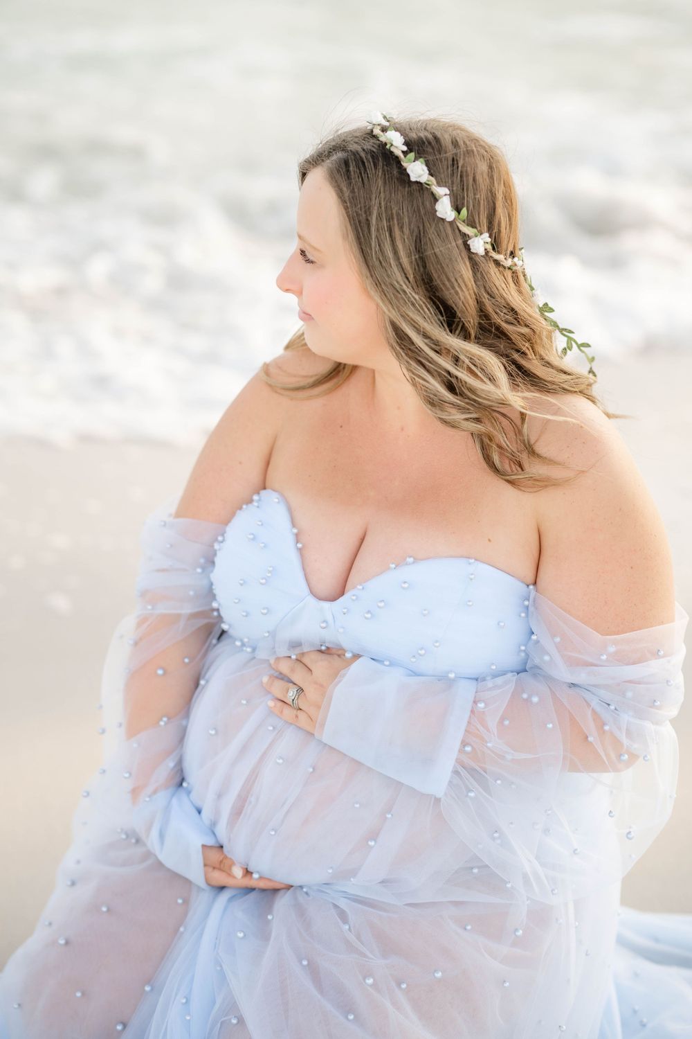 Close-up maternity portrait with delicate floral crown and light blue off-shoulder dress against ocean backdrop.