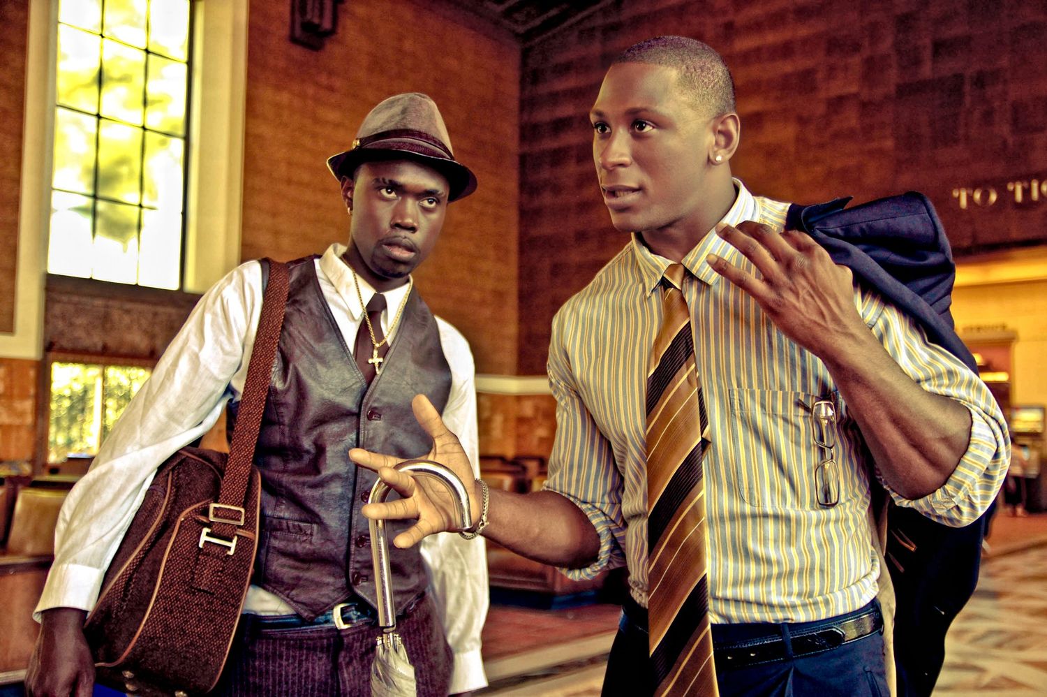 Two men in vintage style clothing having a conversation in a brick-walled room.
