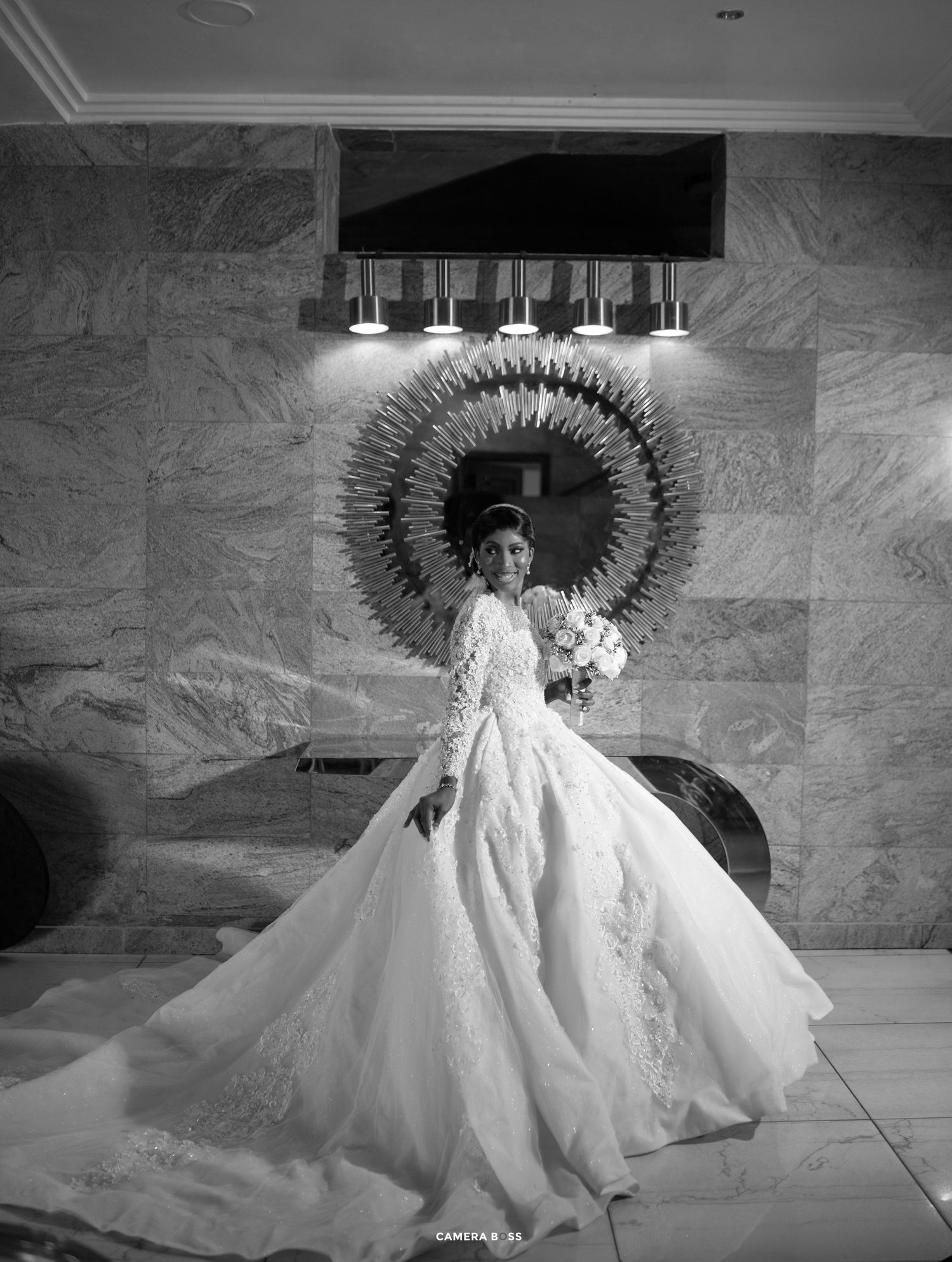Dramatic black and white photo of bride in flowing gown posing by circular wall mirror.