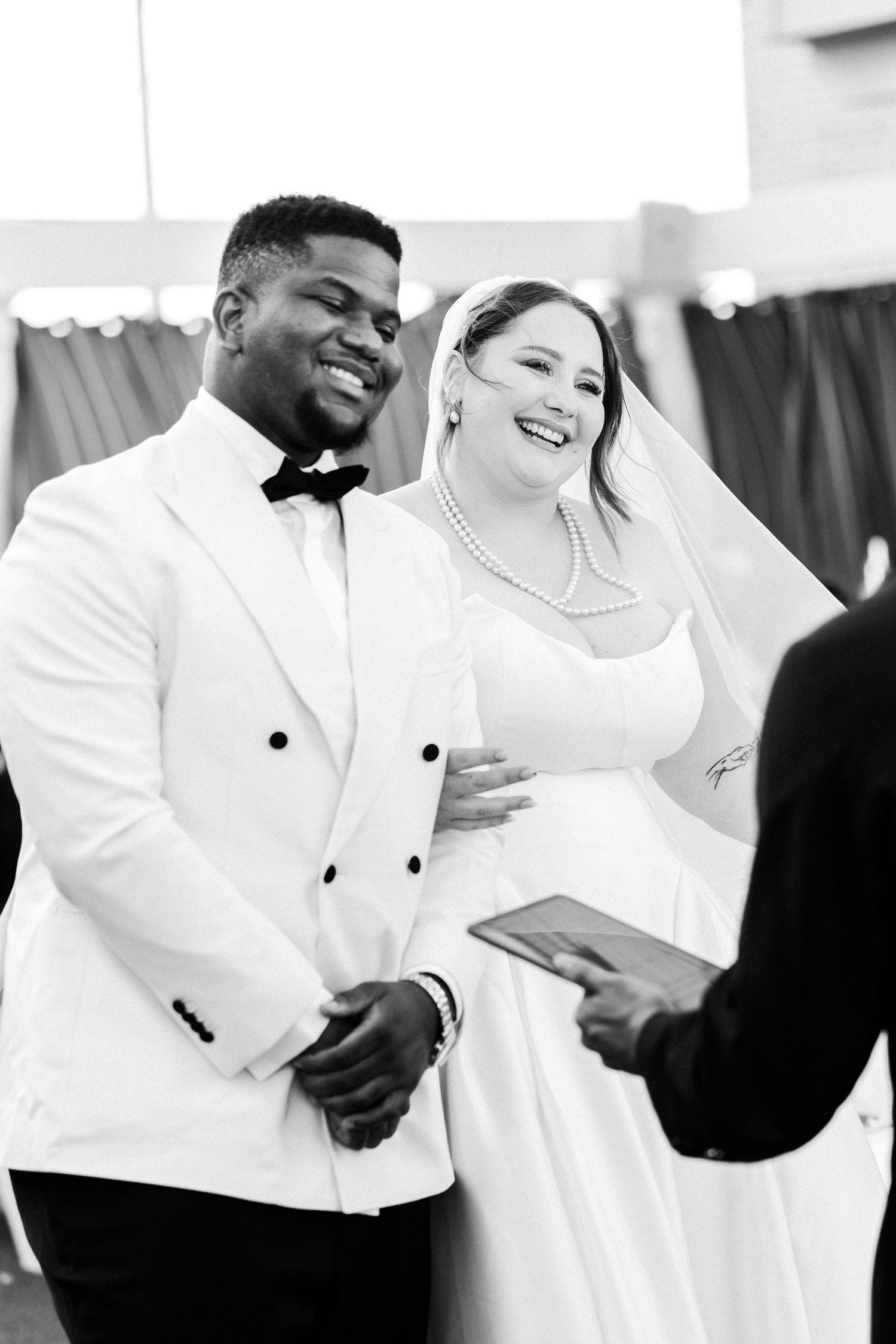Black and white photo of a wedding ring being placed on a bride's finger during ceremony with flowers in background.