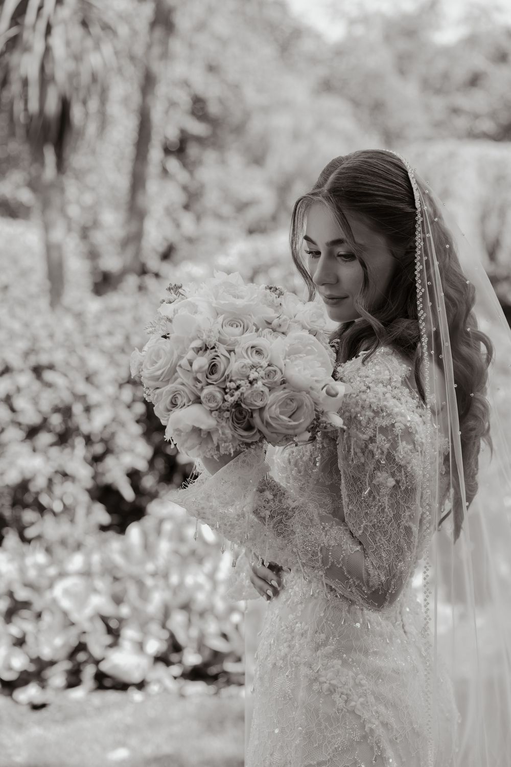 Black and white photo of a bride in lace gown and veil smelling her white rose bouquet outdoors.