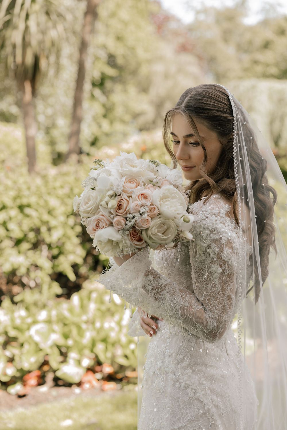 Bride in lace gown and veil holds white and blush rose bouquet outdoors in a lush garden setting.