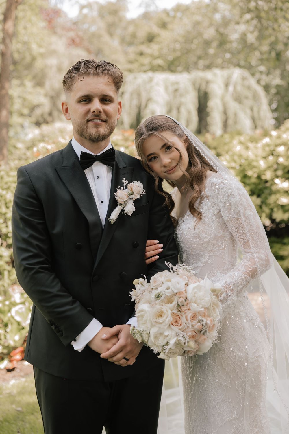 A bride and groom pose together in a lush garden, she holds a white rose bouquet while wearing a lace gown and veil.