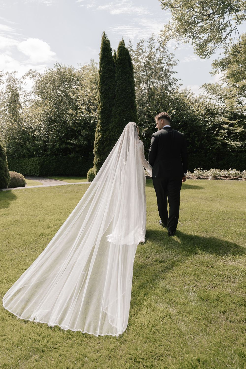 Bride and groom walking hand in hand through a lush garden, bride wearing a long flowing white veil trailing behind.