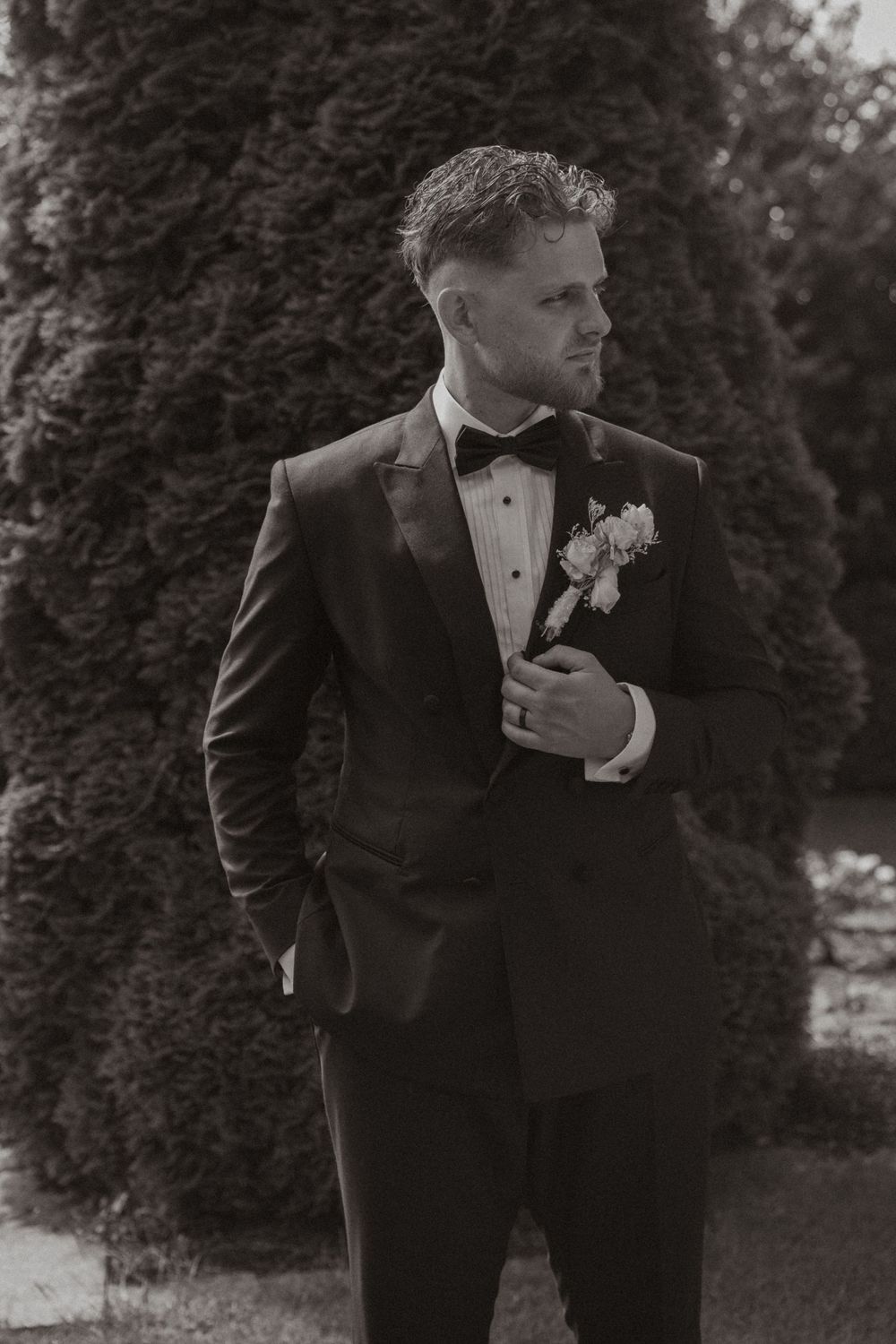 Black and white photo of a groom in a tuxedo with boutonniere, standing outdoors near evergreen trees.