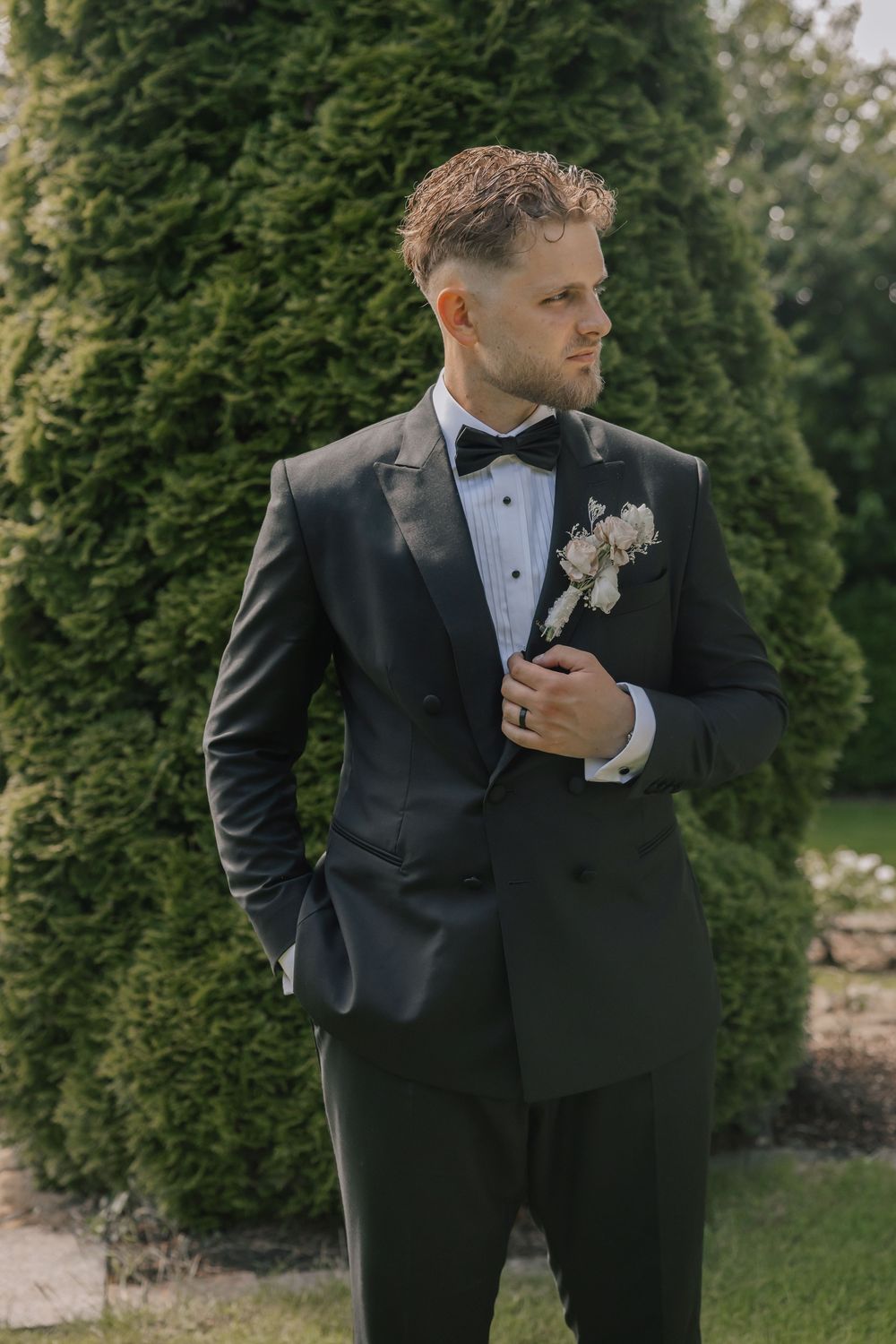 Dapper groom in black tuxedo with bow tie and boutonniere poses outdoors near lush green evergreen trees.