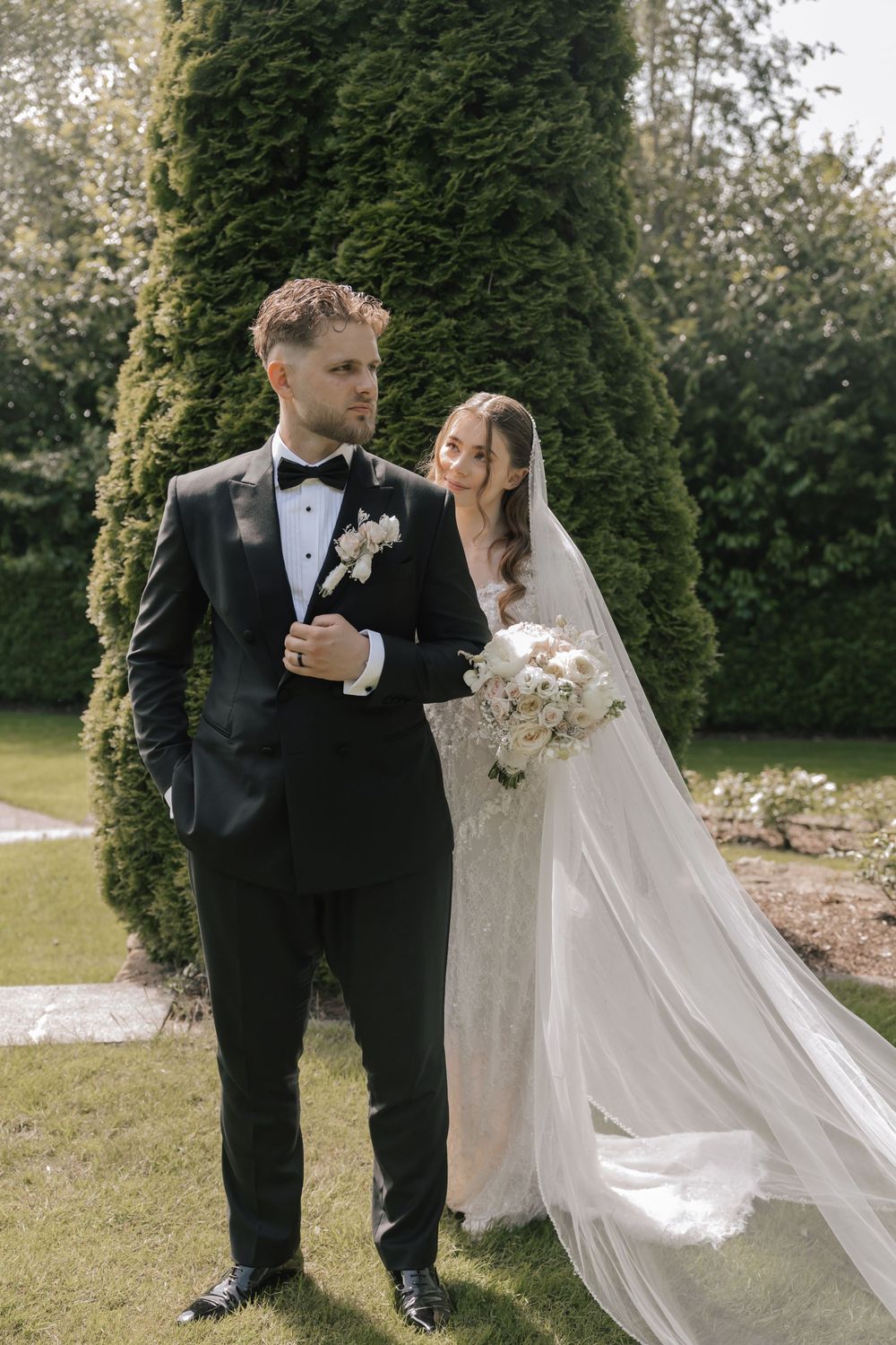 Elegant bride in lace gown with flowing veil stands beside groom in black tuxedo in a formal garden setting.