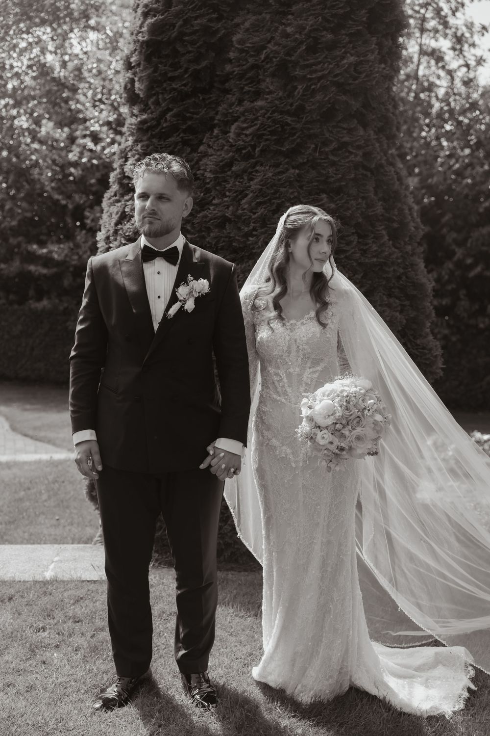 Black and white photo of bride and groom holding hands outdoors, bride wearing lace gown with veil and bouquet.