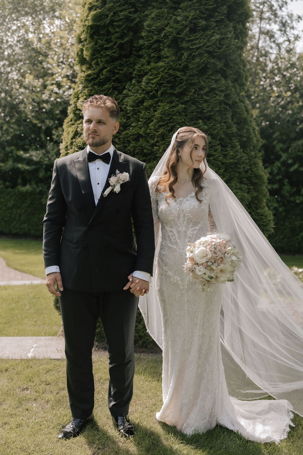 Bride and groom holding hands outdoors, she wears a lace gown with veil, he wears a black tuxedo, green hedges behind.