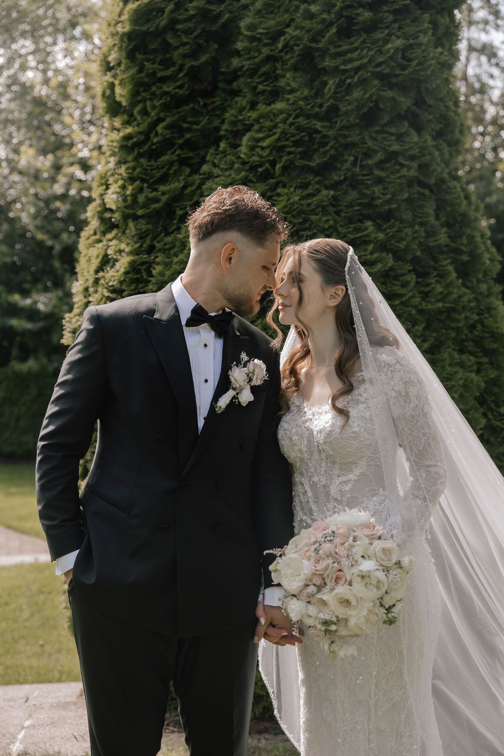 A bride and groom share a romantic moment in a sunlit garden, with the bride holding a white bouquet and wearing a veil.