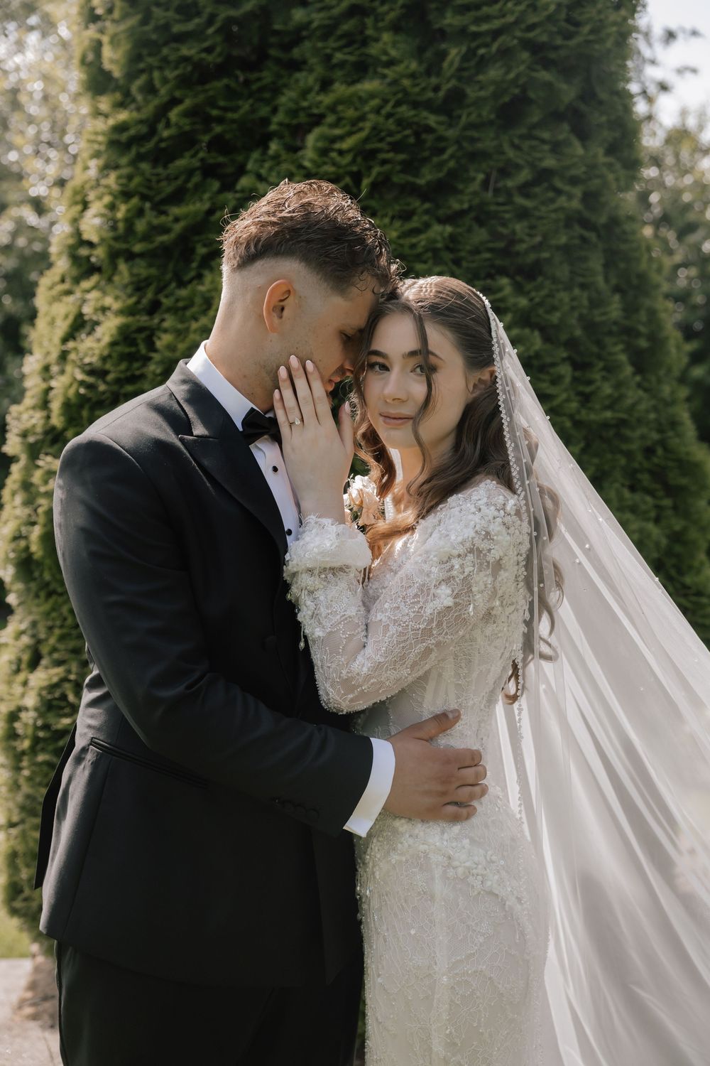 A bride and groom embrace outdoors in front of tall green cypress trees, wearing elegant wedding attire.