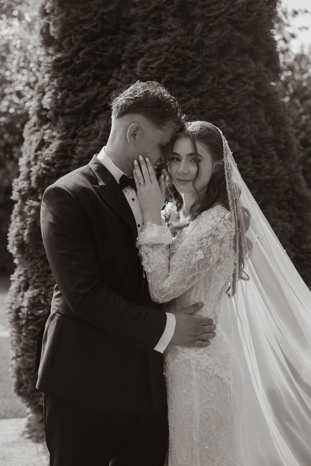 Black and white wedding photo of bride and groom embracing tenderly outdoors near lush dark hedges.