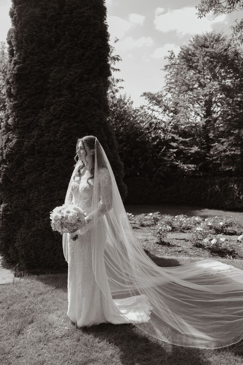 Bride in lace gown with flowing cathedral veil holds bouquet in black and white garden portrait.