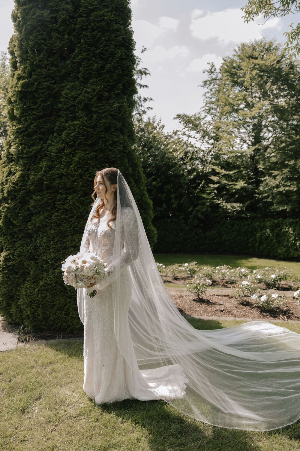 Bride in lace wedding gown with flowing cathedral veil holds white bouquet in lush garden beside tall evergreen trees.