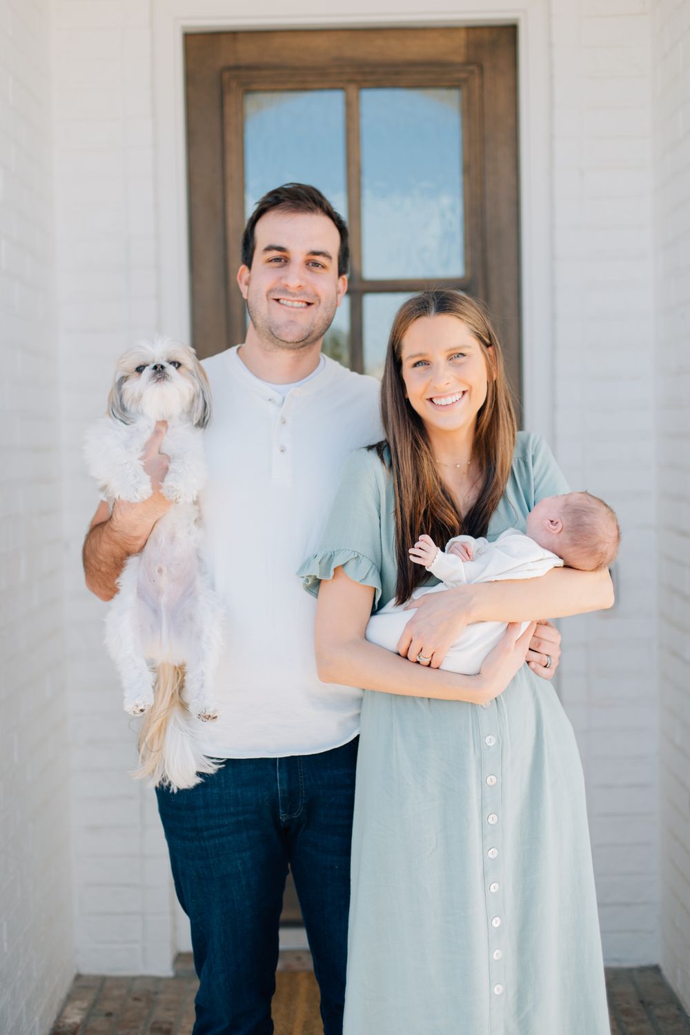 A couple stands outside a white building with a small white dog and newborn baby.