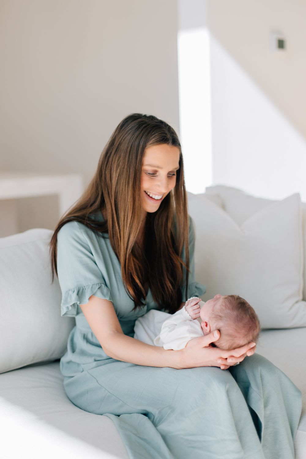 A person in a sage green dress sits on a white couch while holding a smiling baby in their lap.