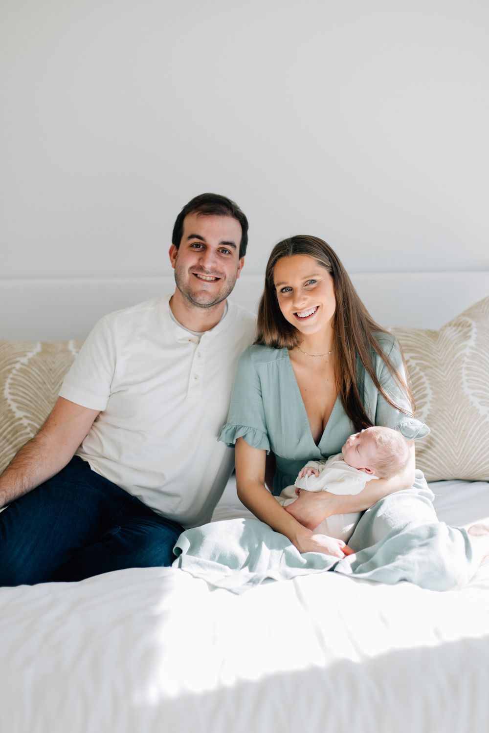 A couple sits on a white bed holding their newborn baby while smiling warmly at the camera.