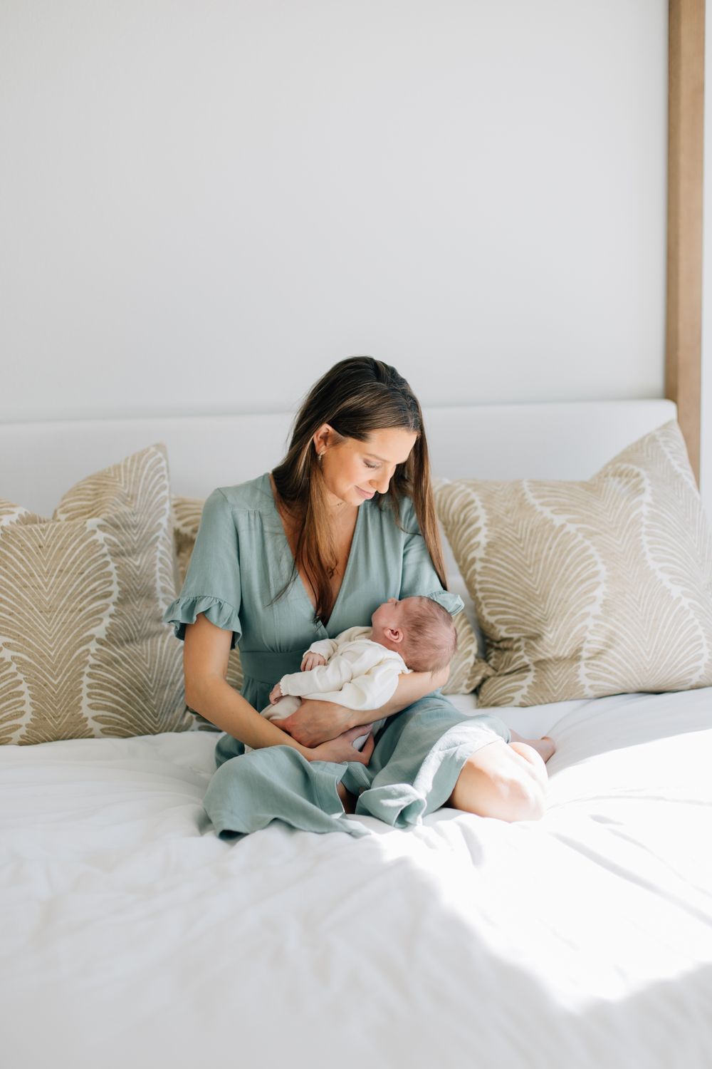 A parent in a sage green dress sits on a white bed with decorative pillows while holding a newborn baby.