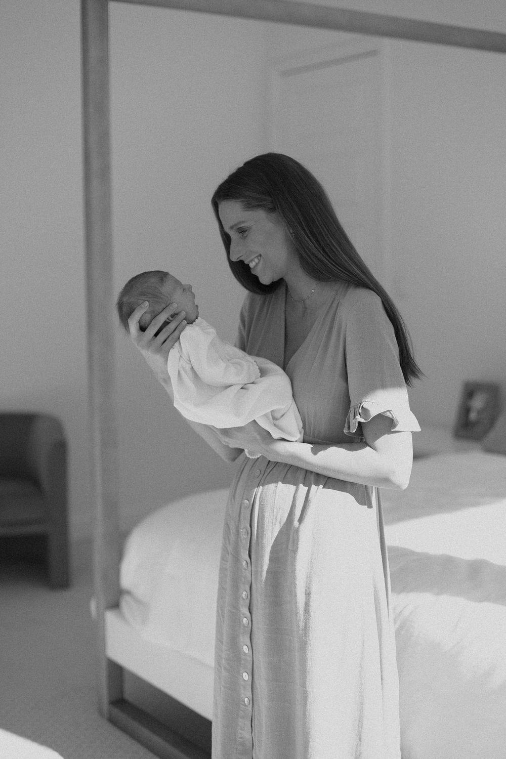A parent in a flowy dress stands near a bedroom mirror while cradling a sleeping newborn baby.