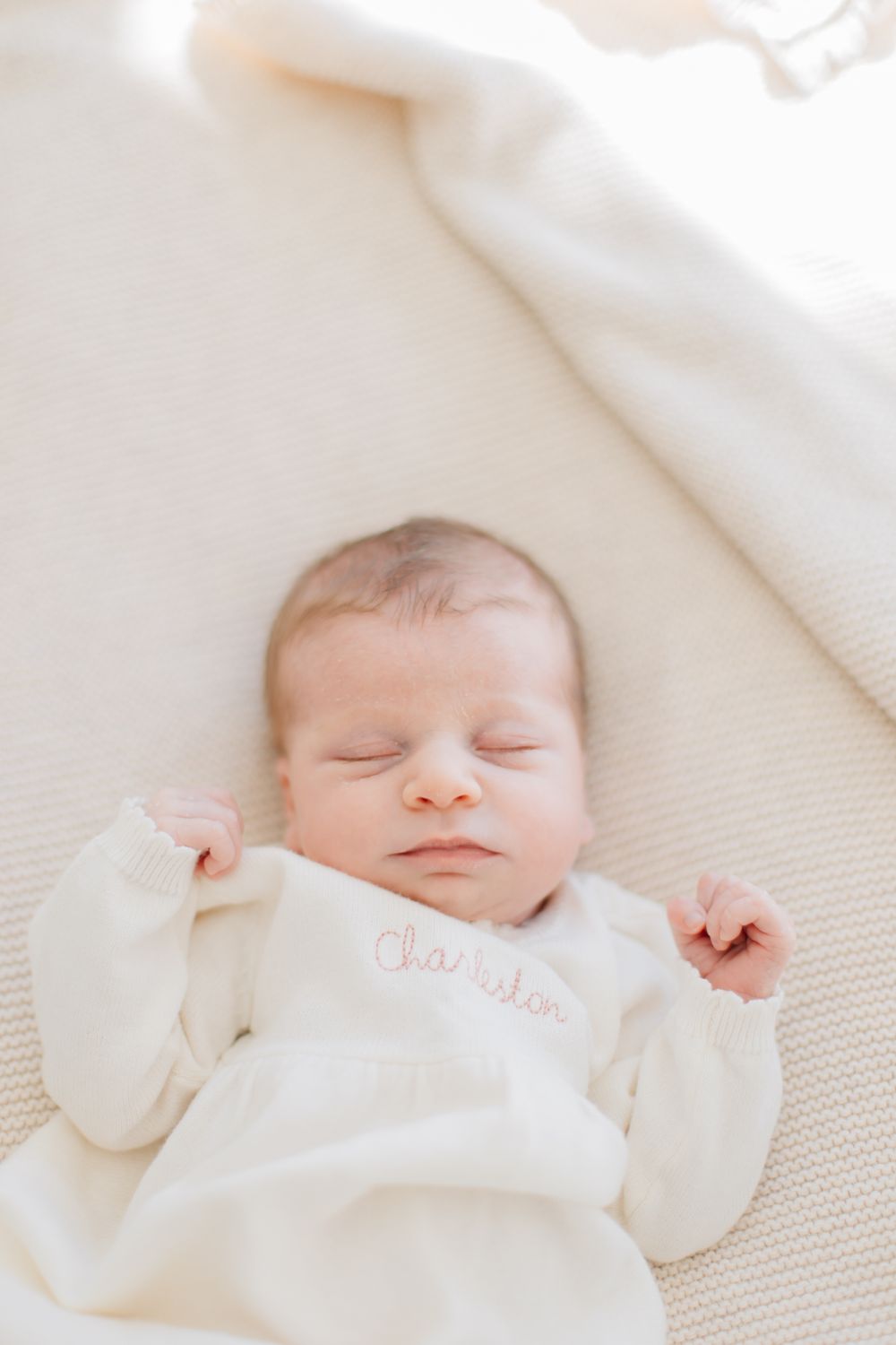 A sleeping newborn baby in a soft cream-colored outfit resting on white bedding.
