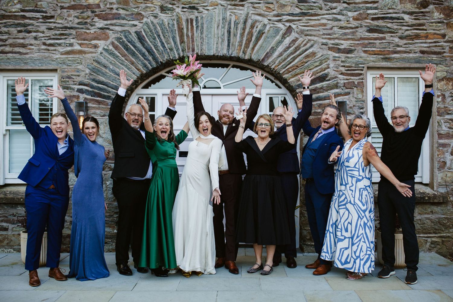 A group of wedding guests pose together under a stone archway, raising their arms in celebration and joy.