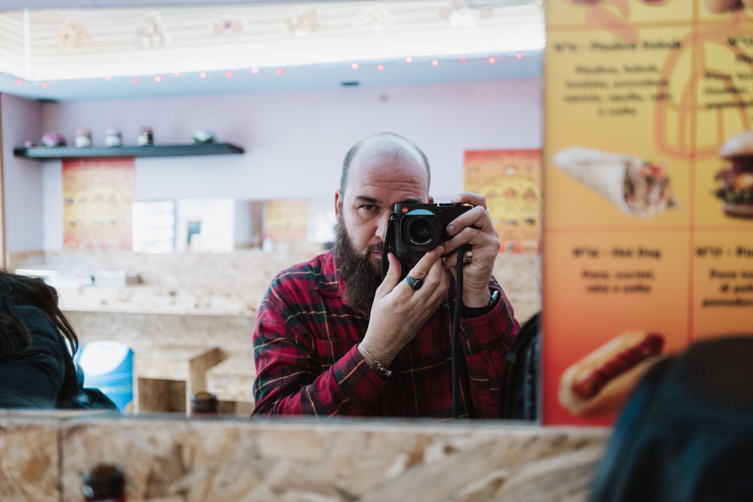 Photographer with beard wearing red plaid shirt takes photo with camera in mirror reflection at cafe.