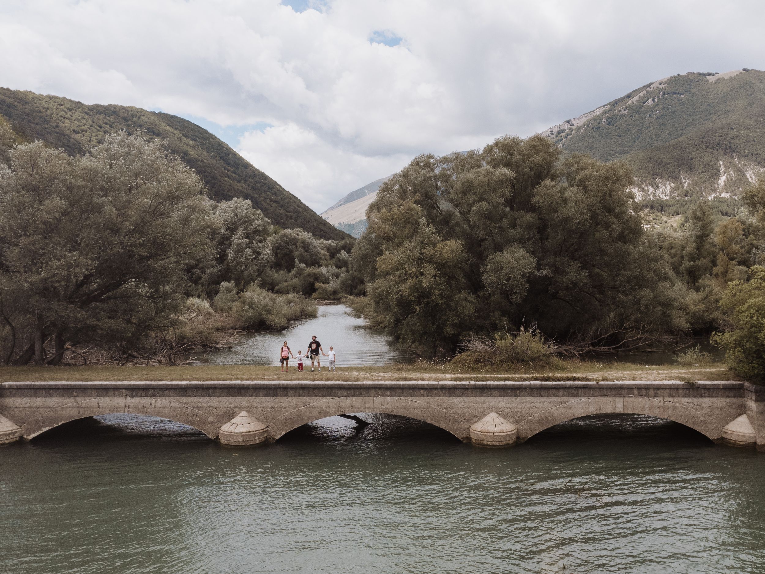 Two people standing on a stone arch bridge over a river surrounded by green mountains and trees.
