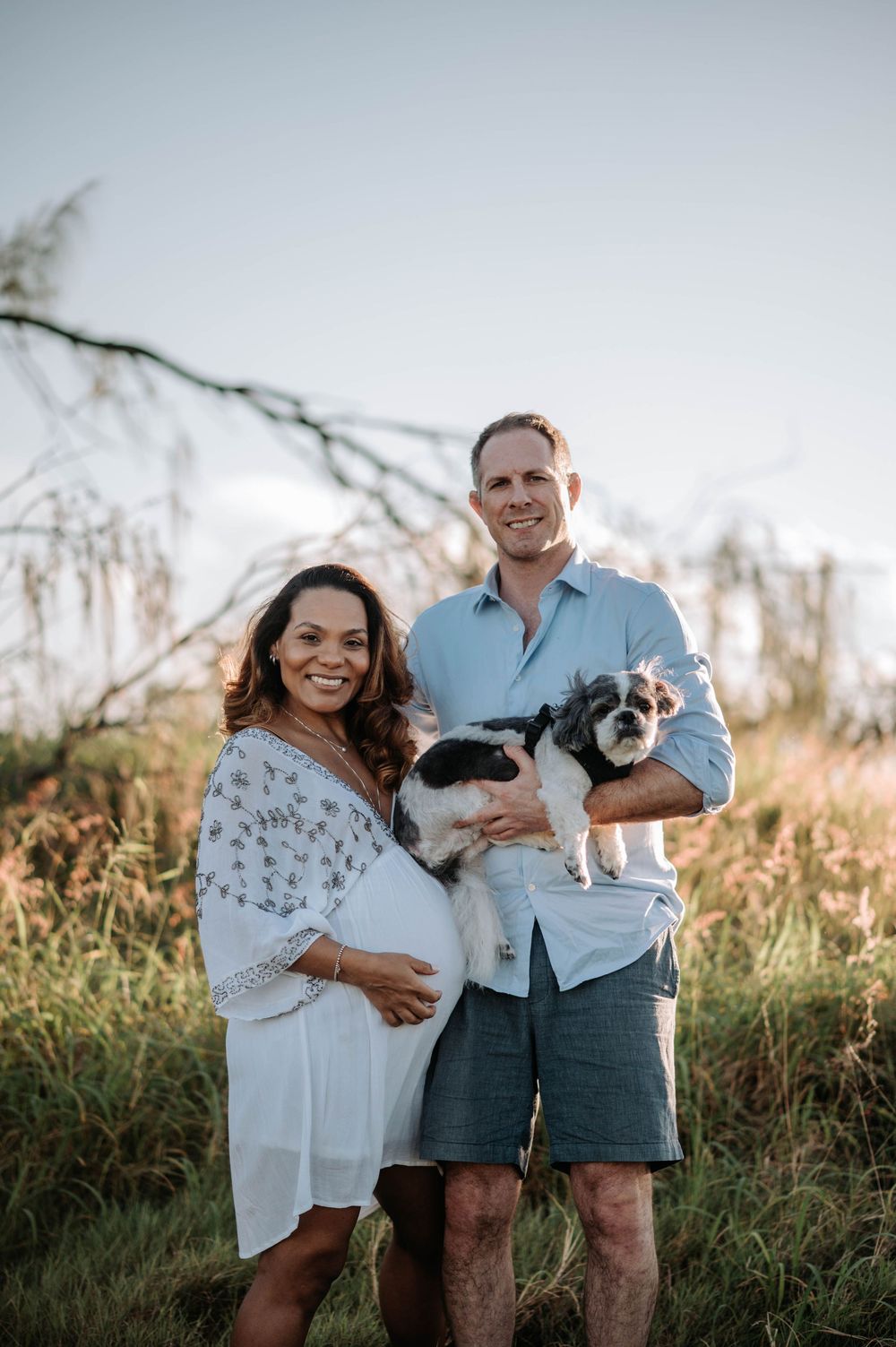 A couple stands together in a field at sunset holding their small black and white dog.