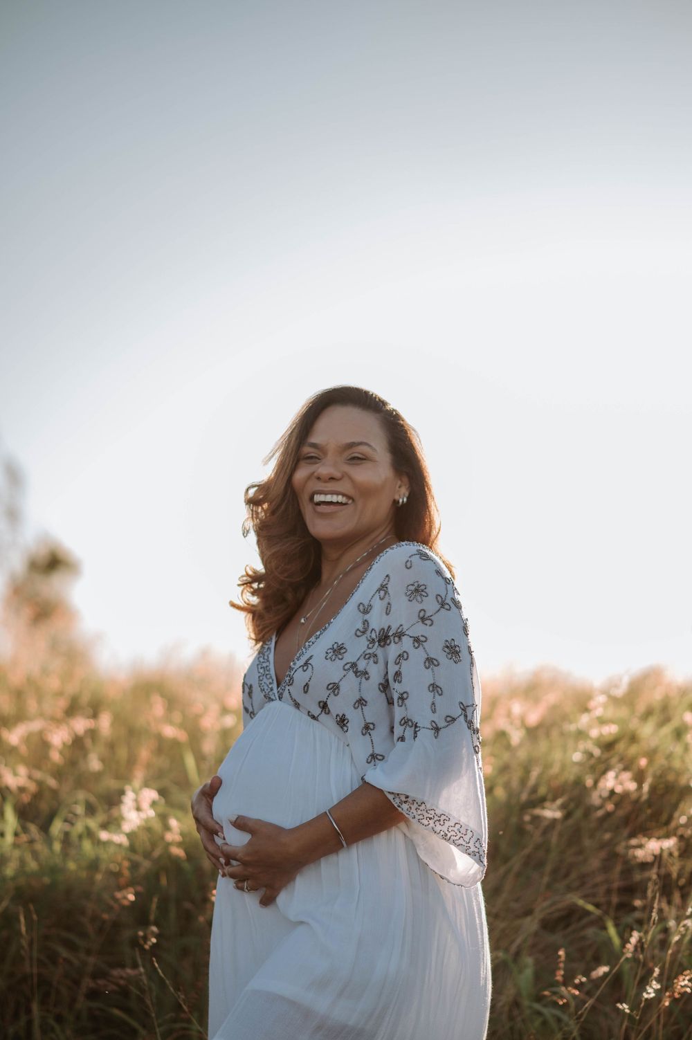 A person wearing a white flowy dress laughs joyfully while standing in a sunlit field with wildflowers.