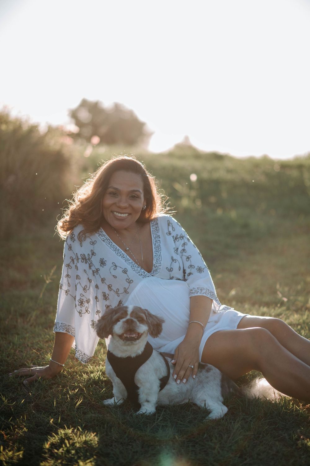 A person in a floral dress sits on grass at sunset with their small black and white dog while golden light streams through.