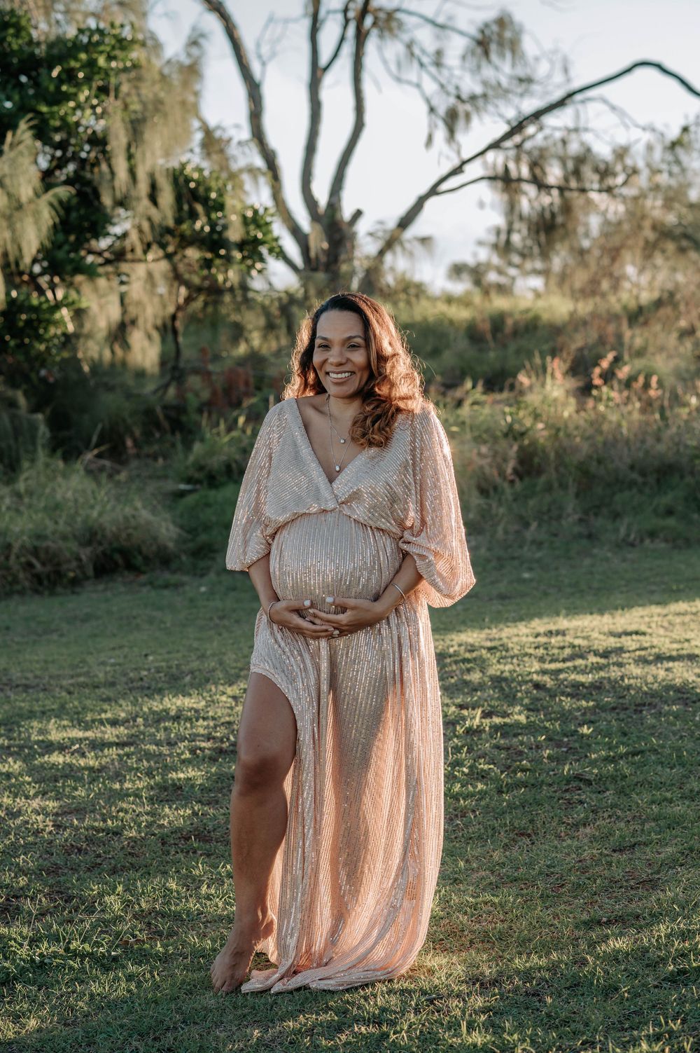 A maternity portrait in a blush sequined gown standing in a sunny garden with Spanish moss draped trees.