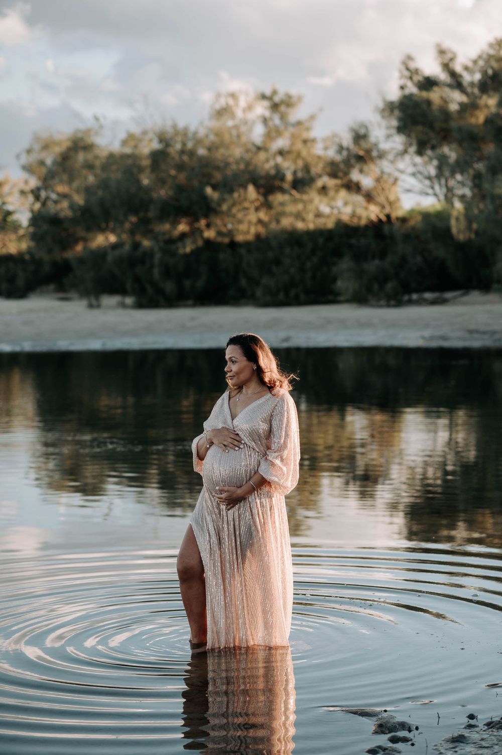 A woman in a flowing white dress stands in calm lake water at sunset creating ripples around her.