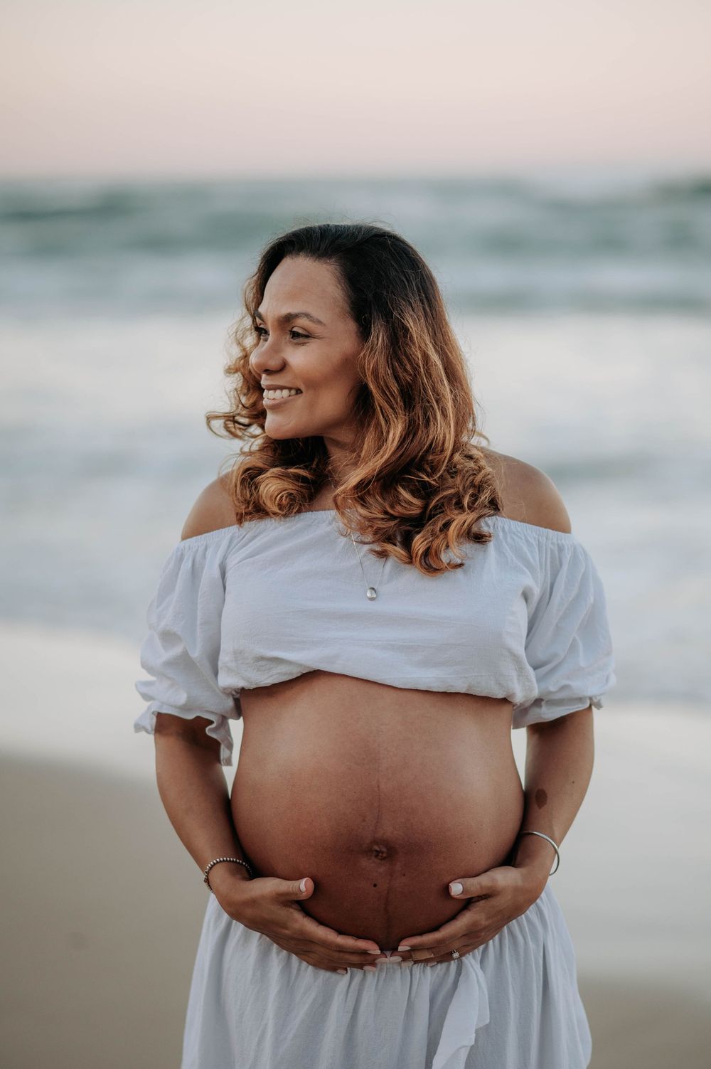 Expectant mother in white off-shoulder dress smiles while cradling her baby bump on beach at sunset.