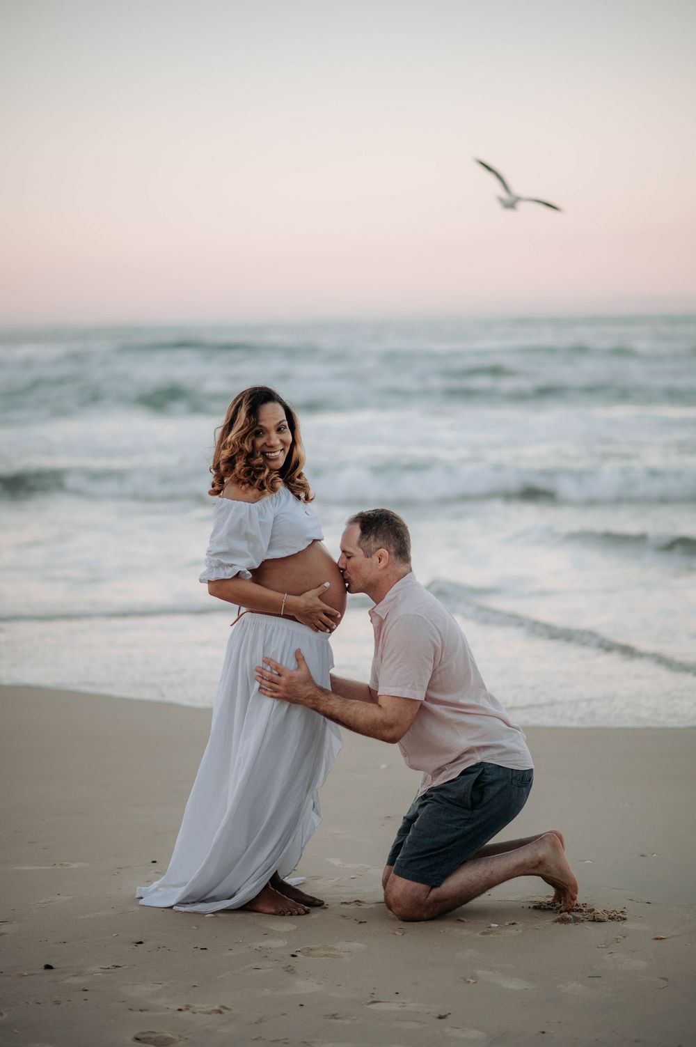 A couple shares a tender moment on the beach at sunset as a seagull flies overhead in the soft pink sky.