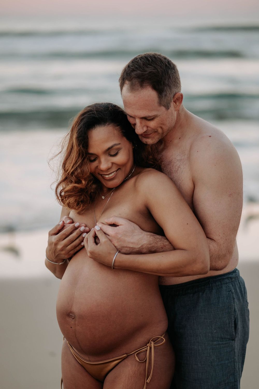 An intimate maternity photo on the beach shows a loving embrace as a couple shares a tender moment during sunset.