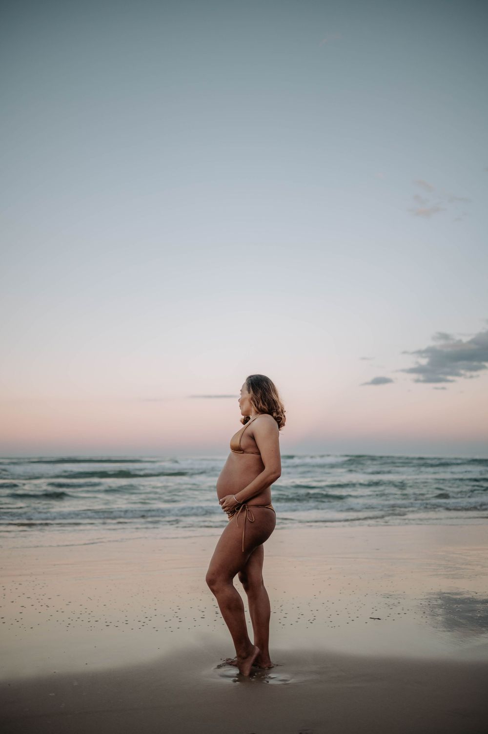 A pregnant figure stands silhouetted against a peaceful beach sunset with gentle waves rolling onto the shore.