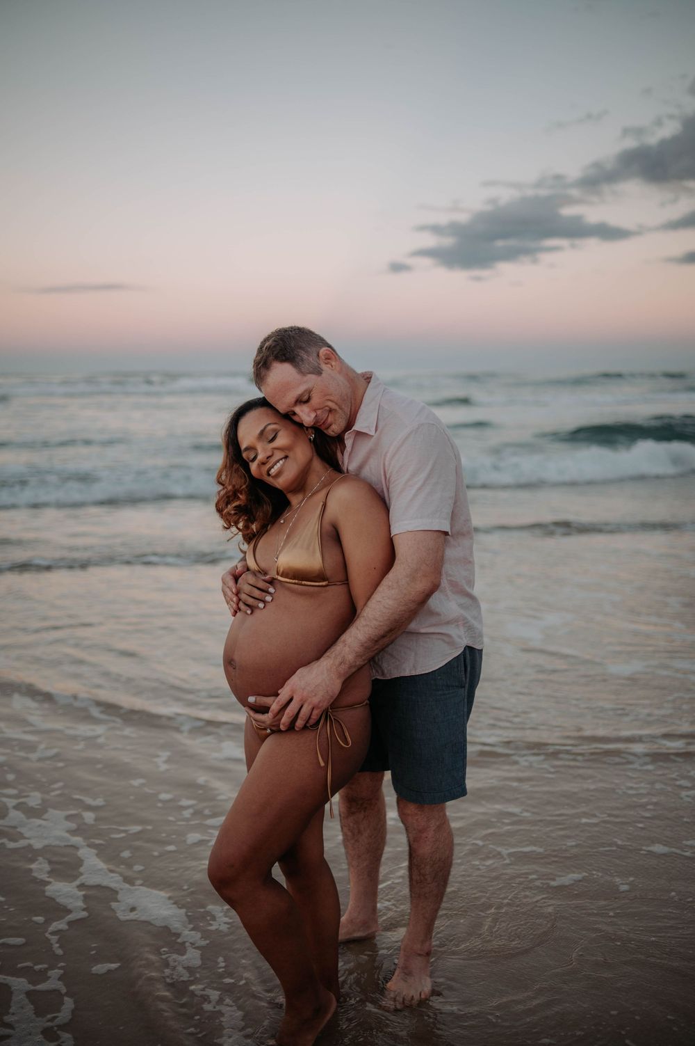 Expectant couple embraces on sandy beach at sunset as gentle waves roll in along the shoreline.