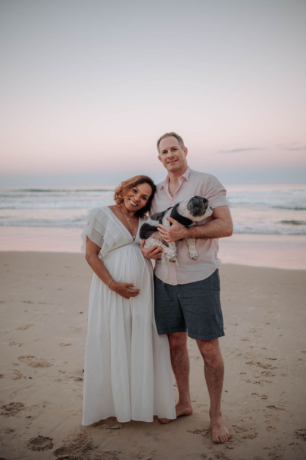 A couple stands on a beach at sunset with their pug dog as waves roll in behind them.